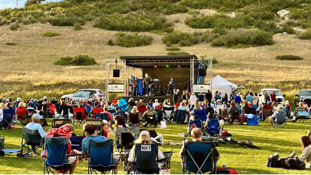 People sitting on the ground and in lawn chairs watching an outdoor summer concert.