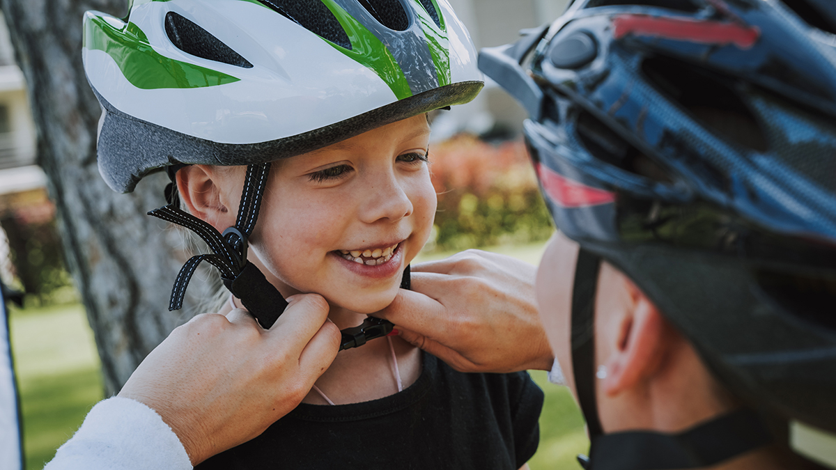 Cheerful little girl looking at mom and smiling while she fastening her helmet