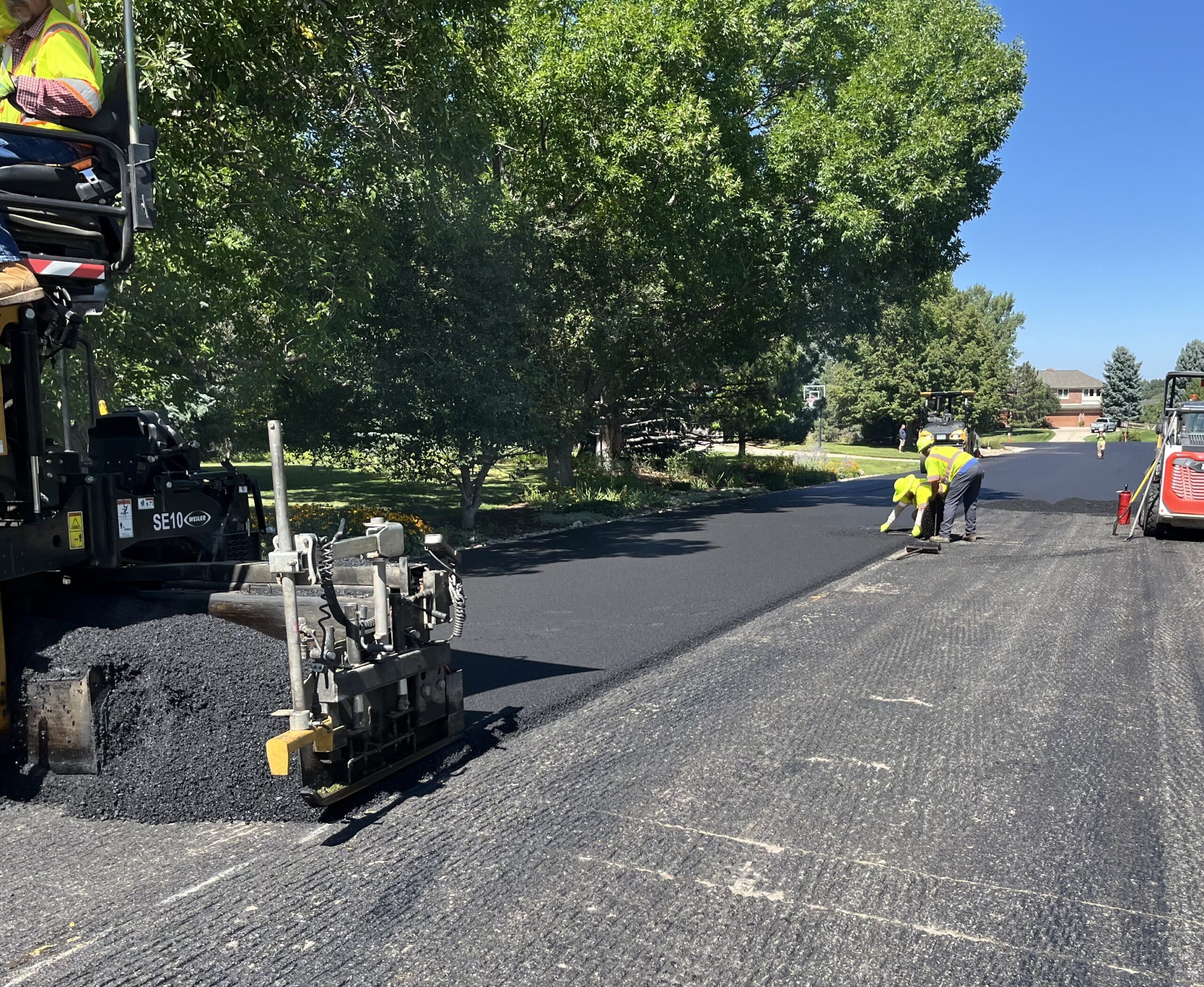 Three people wearing yellow safety vests working on a neighborhood road. Half of the road has a new top layer and the other does not.