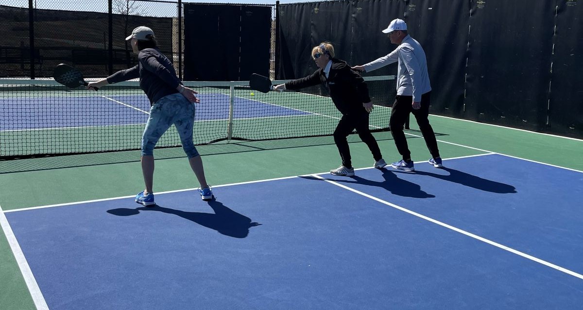 Three people on an outdoor pickleball court holding paddles and working on technique.